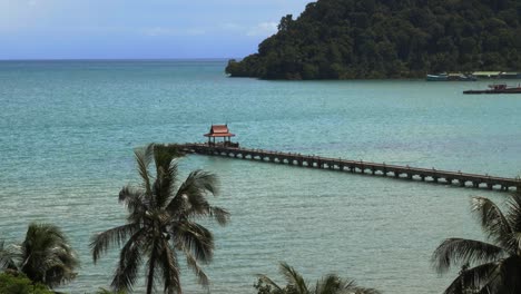 Un-Muelle-Con-Un-Pequeño-Techo-Rojo-Al-Final,-En-El-Océano-Con-Palmeras-A-Cámara-Lenta