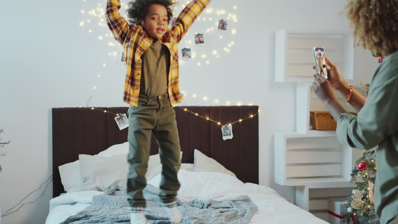 Kid Jumping in Bedroom Decorated for Christmas, Mom Taking Video with Phone