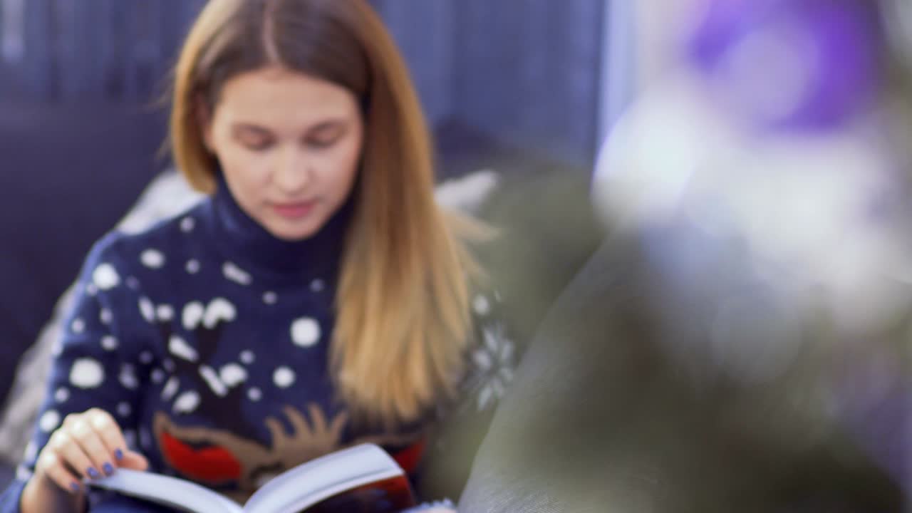 hermosa chica en suéter leyendo el libro sentada cerca del árbol de navidad en el sofá