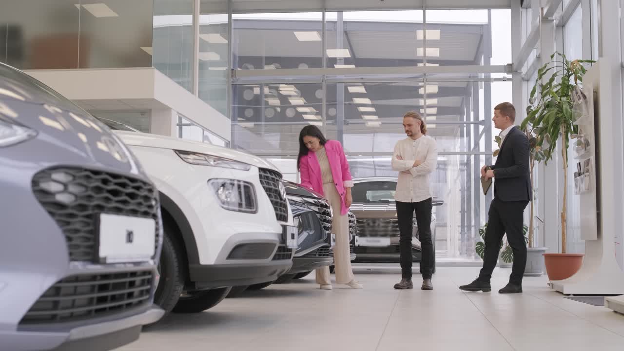 un joven vendedor de coches mostrando a una pareja de jóvenes un nuevo automóvil en el salón de concesionarios.
