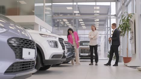 young car salesman showing to young couple new automobile at dealership salon.