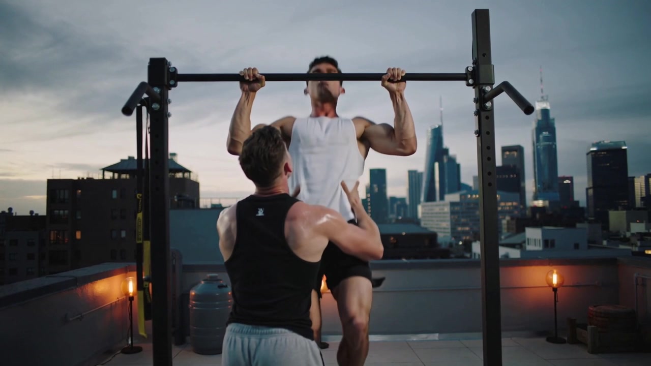 Spotter placing hands for safety while exerciser pulling up on rooftop rig in athletic wear