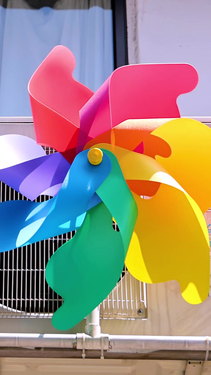 Un molinillo de viento vibrante y multicolor gira rápidamente frente a una ventana y una rejilla metálica, iluminado por una fuerte luz natural del día con un ángulo de cámara estático.