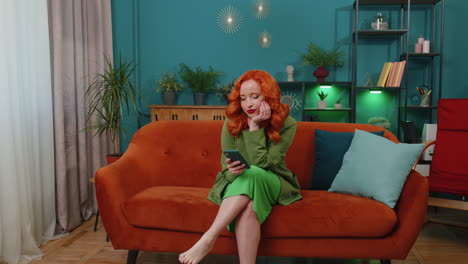 cheerful redhead girl sitting on sofa, using smartphone share messages on social media application