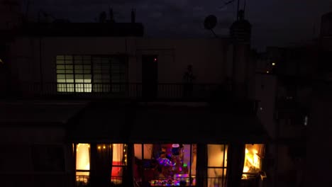 Aerial-view-dolly-out-tilt-down-group-of-friends-celebrating-in-a-building-with-colored-lights-and-yellow-bulbs,-city-at-night