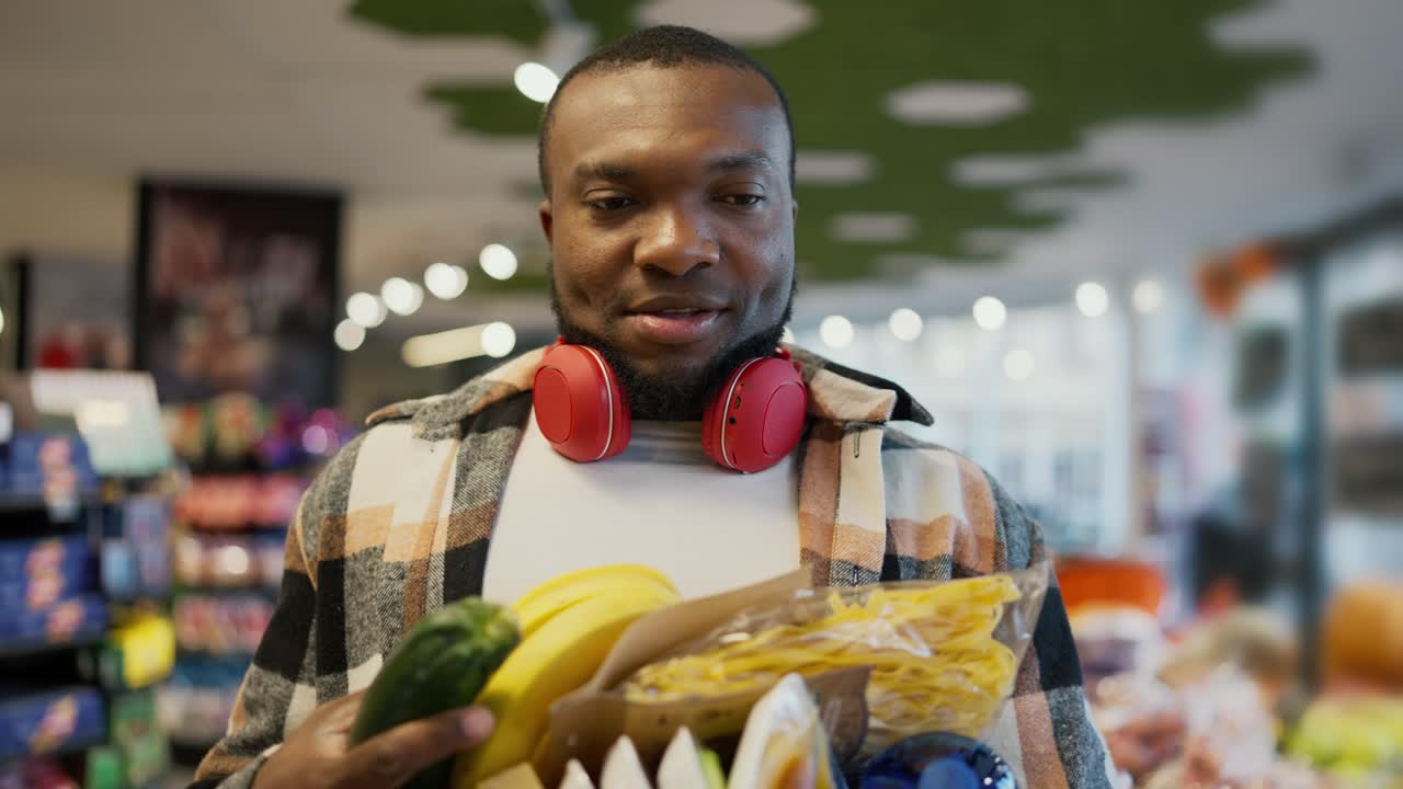 un hombre con piel negra en una camisa a cuadros y usando auriculares inalámbricos rojos difícilmente puede llevar una gran cantidad de comida en su pecho con ambas manos. el hombre no usa una bolsa, pero lleva una gran cantidad de comestibles en su peito sosteniéndolos con la mano