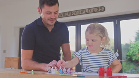 Father-and-young-daughter-having-fun-at-home-sitting-at-table-and-painting-decoration-together---shot-in-slow-motion