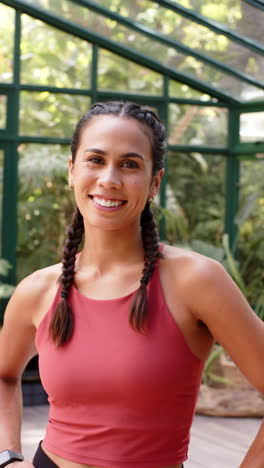 vertical video: smiling woman in yoga attire standing in glass house, ready for yoga session