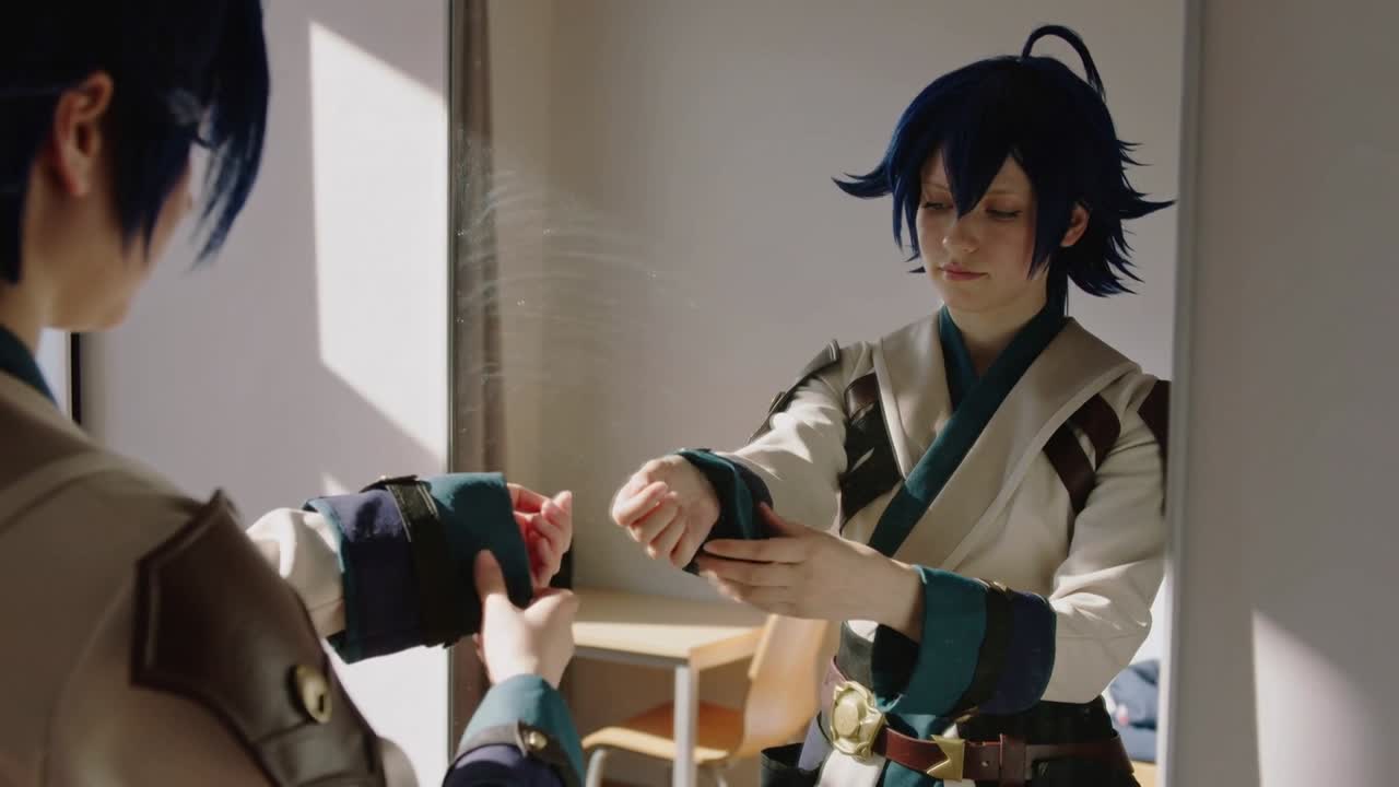 Đối diện gương, cosplayer mặc áo choàng và áo giáp màu xanh mòng két đang điều chỉnh còng tay trong căn phòng ngập nắng, chuẩn bị trang bị.