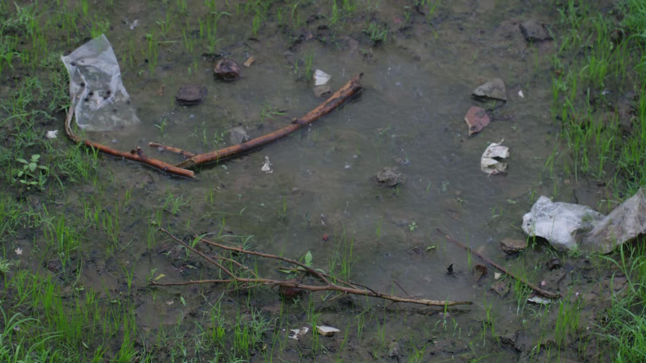 A top angle handheld mid shot of a puddle of rainwater, plastic and wooden sticks on ground while it rains heavily