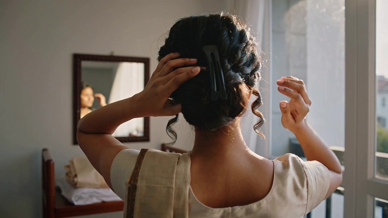 Raising hands woman in sari securing bun at balcony using large black clip checking mirror prepping