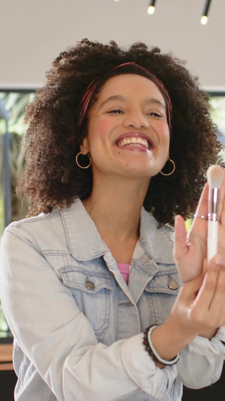 Vertical video: Camera focusing, woman in denim jacket demoing makeup brush at home table