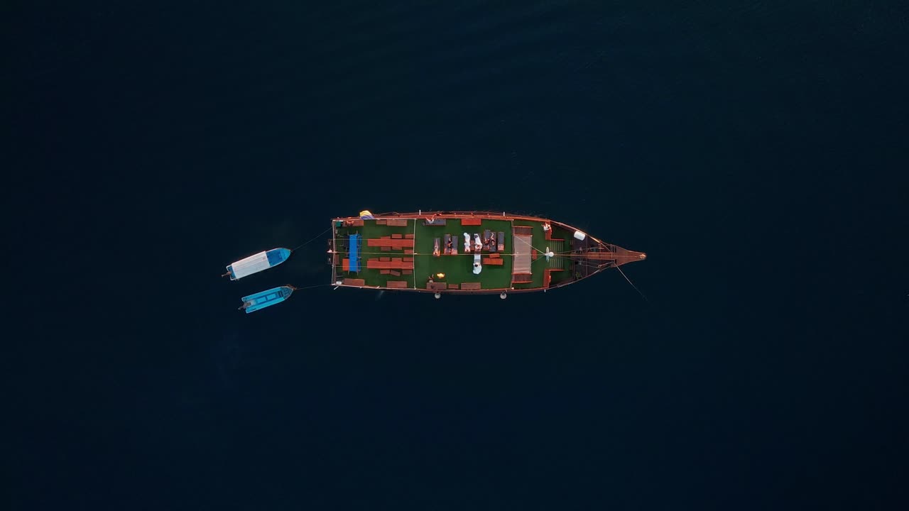 A zoomed-out overhead view shows a large Phinisi tour boat sailing through the deep, dark blue ocean, with two small boats traveling right beside it.