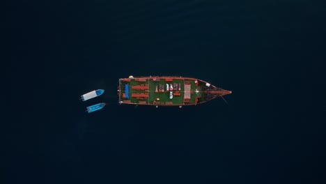 a zoomed-out overhead view shows a large phinisi tour boat sailing through the deep, dark blue ocean, with two small boats traveling right beside it.
