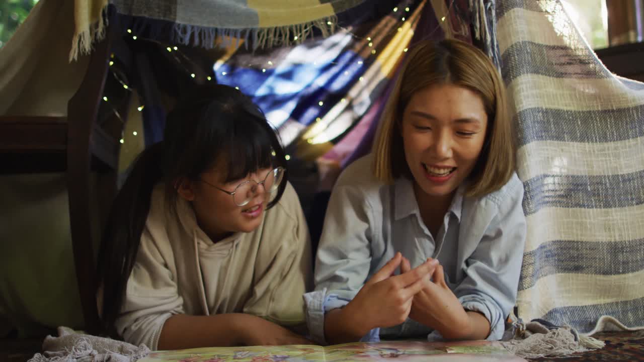 아시아 어머니 가 책 을 읽고 집 에서 담요 요새  ⁇  에 딸 을 안아주고 있다