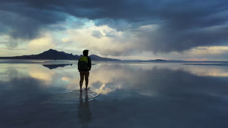 silhouette of man with backpack walking in surreal landscape of salt flats with stormy clouds and sky mirror reflection on water, cinematic approaching view