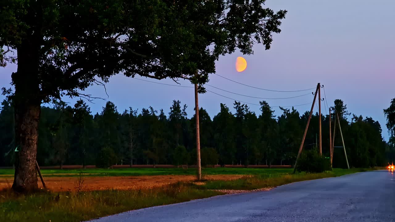 Quiet rural road with rising moon above trees and power lines during blue evening sky