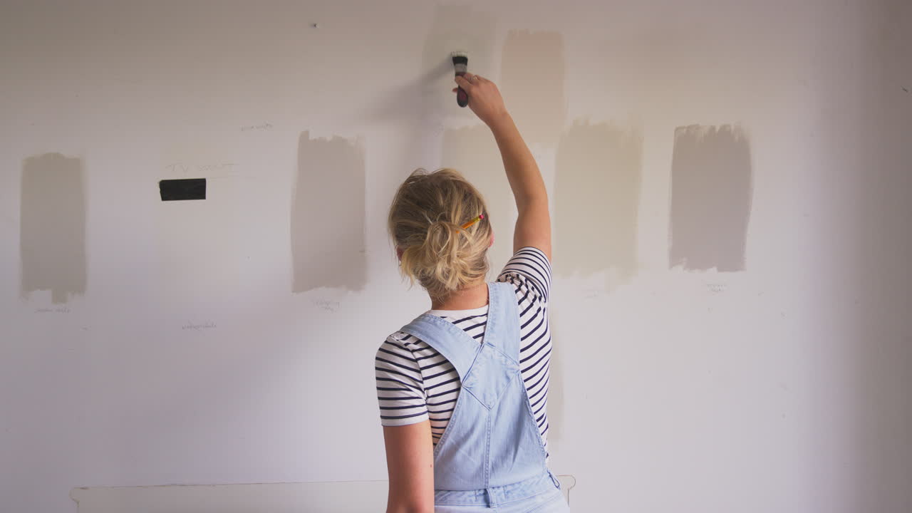 Rear View Of Woman Decorating Room In House Painting Tester Paint Colour Strips On Wall