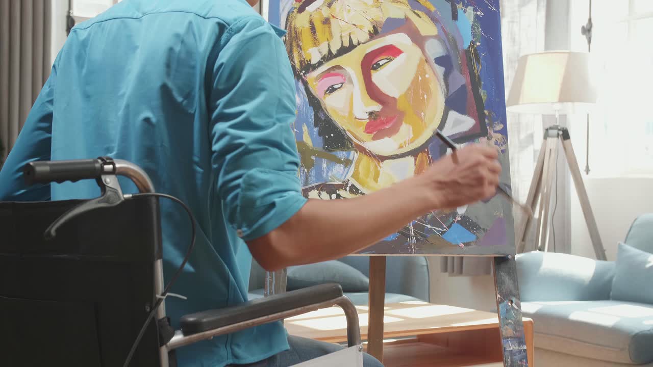 Medium Close Up Hind View Of An Asian Artist Man In Wheelchair Holding Paintbrush And Mixing It With Colour Before Painting A Girl'S Face On The Canvas