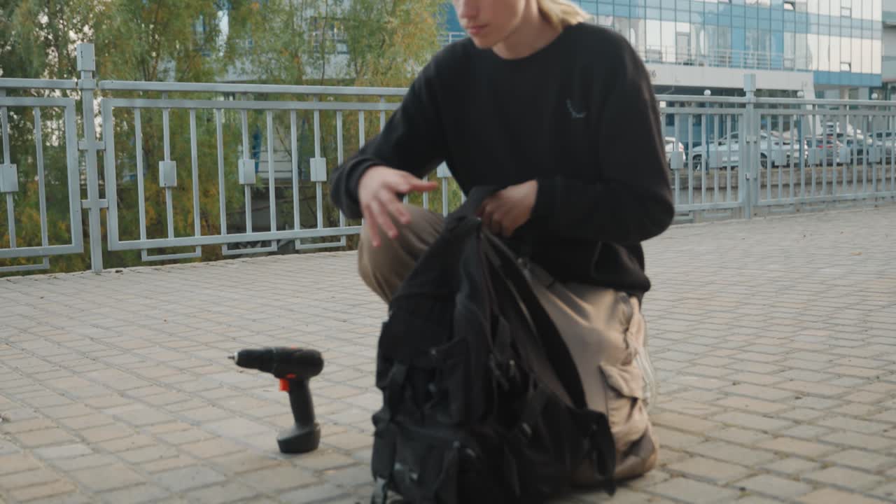Esploratore urbano caucasico che disfa l'attrezzatura vicino a un moderno lungomare, camminando tra edifici di vetro, inginocchiato per aprire lo zaino, controllo metodico dell'attrezzatura, attrezzo manuale e caricabatterie visibili, paesaggio urbano diurno