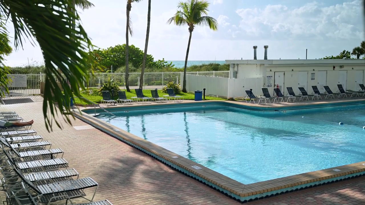 Udendørs swimmingpool med palmetræer nær havet, Miami Beach, Florida. Murstensdæk med liggestole, grøn græsplæne og hvidt hegn under delvist overskyet himmel.