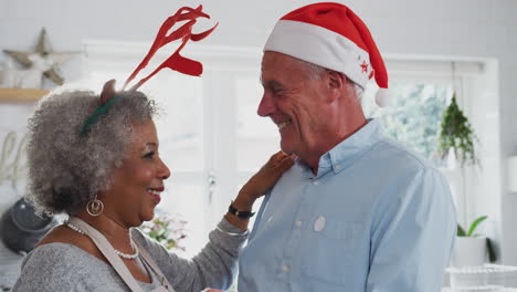 Loving-Senior-Couple-Wearing-Fancy-Dress-Antlers-Dance-In-Kitchen-Whilst-Preparing-Christmas-Dinner