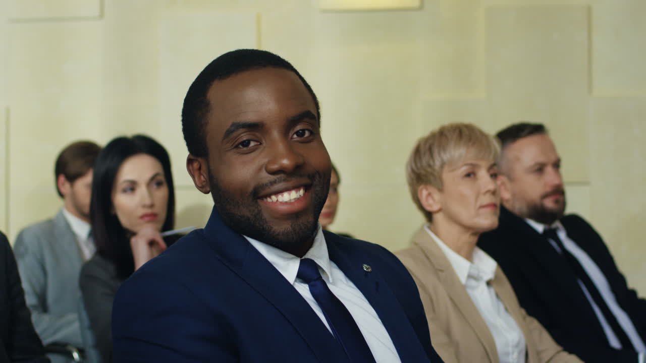 hombre de negocios afroamericano sentado en una silla entre personas en una sala de conferencias sonriendo y mirando la cámara