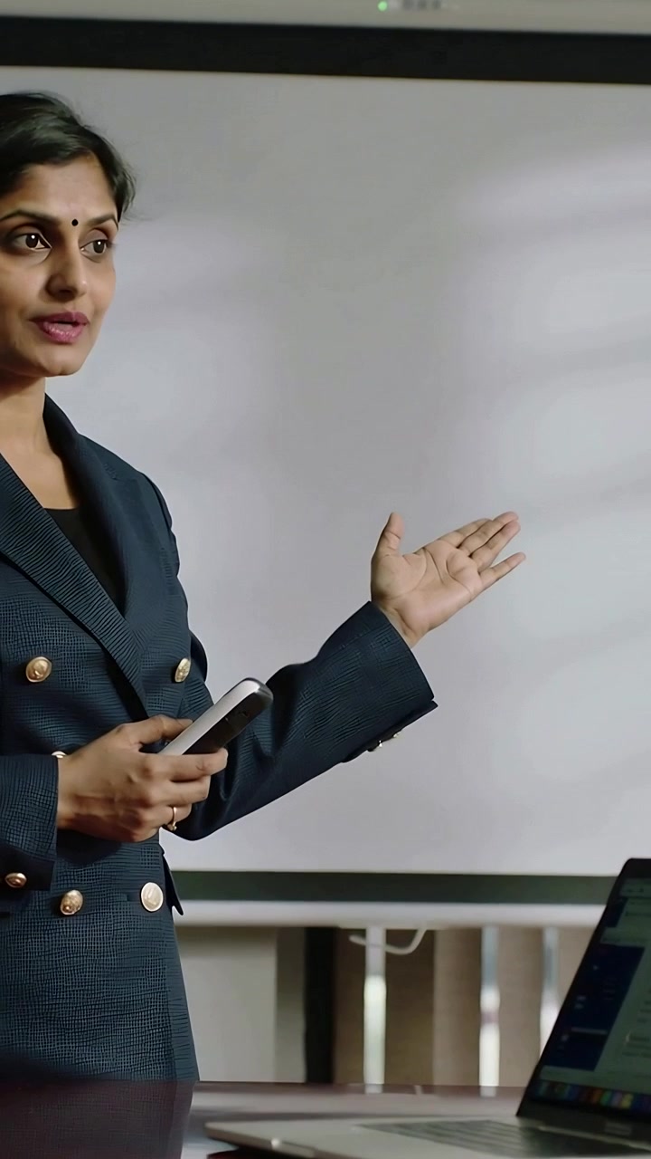 Video verticale: Donna in tailleur che usa il telefono e indica punti sullo schermo e sul laptop in ufficio.