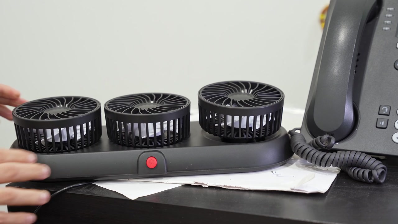 Hands adjust three portable desk fans on a shared base beside a landline phone, demonstrating personal cooling equipment in a modern office interior.