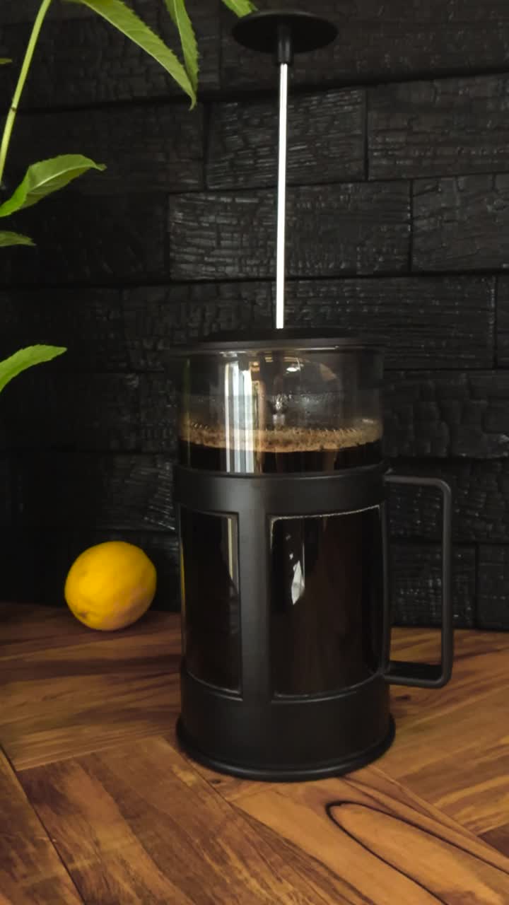 Gorgeous close up or closeup footage of a dark coffee filled prench press on a olive wood table that has a lemon and peppermint plant on it and a dark black Yakisugi burnt wooden wall in the back.