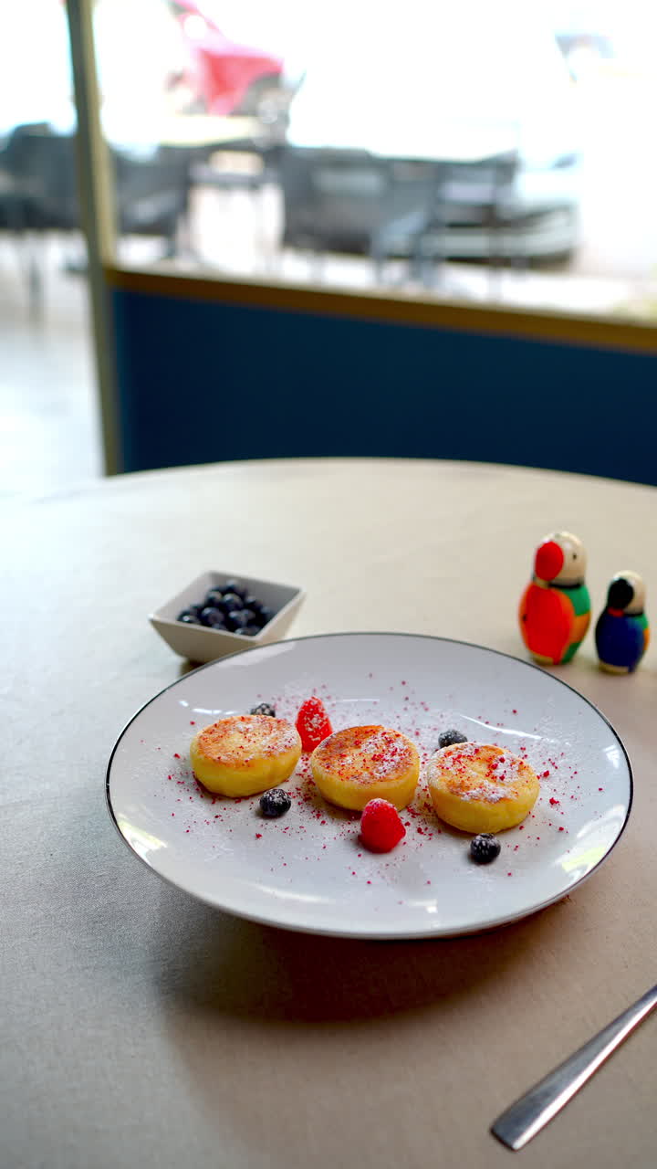 Deliziosi syrniki, tipici pancake di formaggio dell'Europa orientale, serviti su un piatto bianco e decorati con lamponi, mirtilli e zucchero a velo, perfetti per la colazione o il brunch.