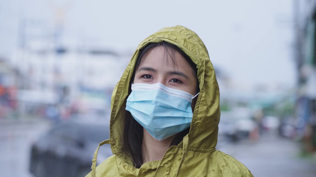una joven y bonita chica asiática con máscara protectora, impermeable amarillo de pie en el lado de la carretera, con el coche estacionado en la calle urbana en el fondo, mantenerse a salvo en la ciudad contaminada, la temporada de lluvias clima