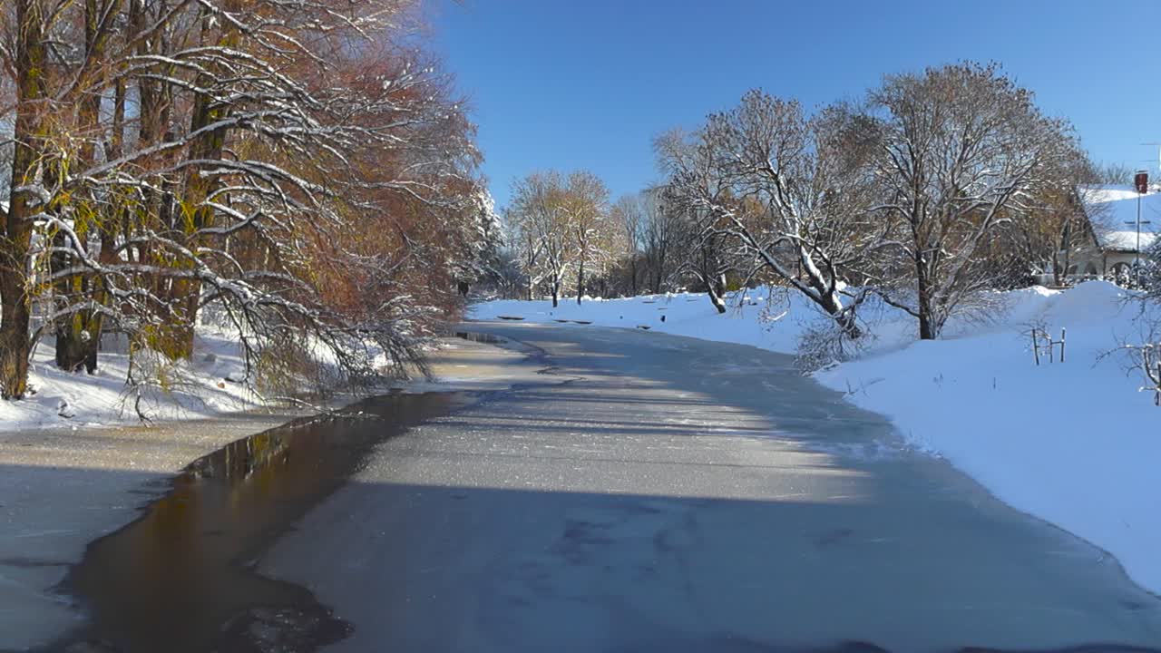 Gorgeous footage of a river in Laagri slightly covered with ice and snow while big brown oak and ash trees grown on the riverbanks during sunny winter day. Trees and riverbanks covered in white snow.