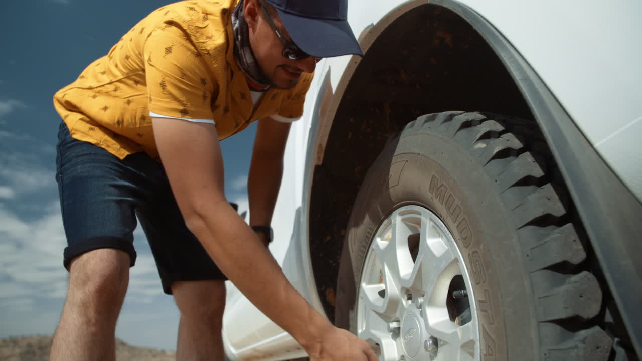 Low angle wide hand held shot of a Caucasian male tourist in Africa as he removes loose a lug nut by hand from a flat tire