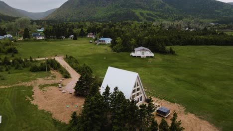 Aerial-drone-shot-panning-downward-of-a-beautiful-scene-with-the-Cape-Breton-Highlands-in-the-background-revealing-an-A-Frame-cabin-off-the-coast-of-the-Atlantic-Ocean-in-Nova-Scotia