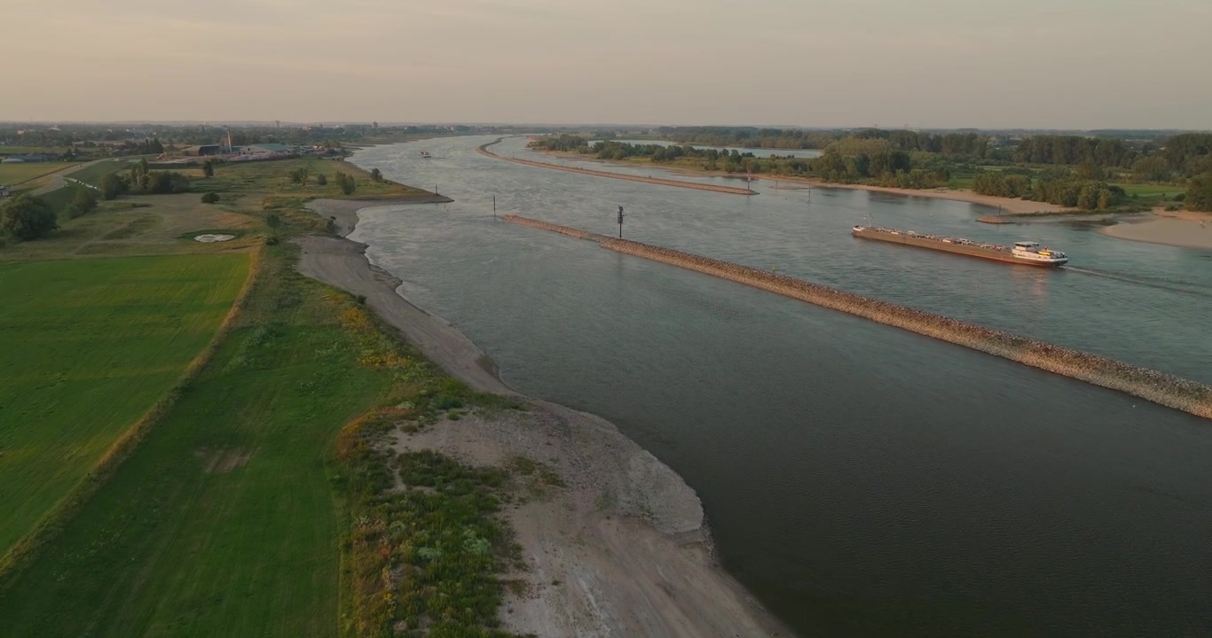 Un drone si avvicina a una nave da carico dalla riva del fiume, rivelando un fiume tortuoso, argini protettivi e l'ampio paesaggio dei polder olandesi, mettendo in mostra la navigazione interna e la gestione dell'acqua