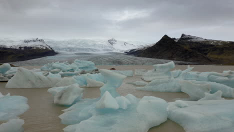 Aerial:-Flying-low-over-icebergs-floating-in-the-lagoon-and-a-small-boat-with-tourists-in-Fjallsarlon-glacier-in-Iceland-during-a-cloudy-day