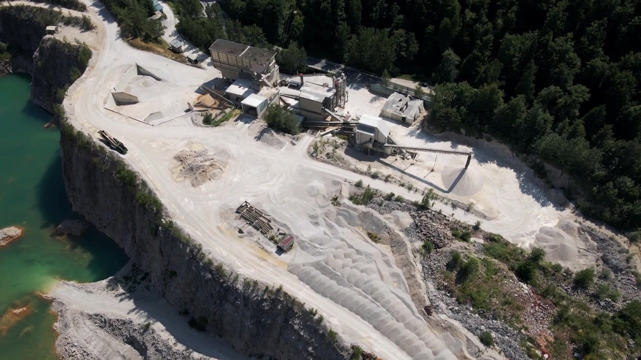 Weitläufige Luftaufnahmen eines Kalksteinbruchs neben türkisfarbenem Wasser in Savinjska, Slowenien, enthüllen dramatische Klippen, Maschinen und einen üppigen Waldrand