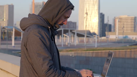 Cold-hooded-male-hacker-quickly-typing-code-on-laptop-overlooking-downtown-Barcelona-waterfront