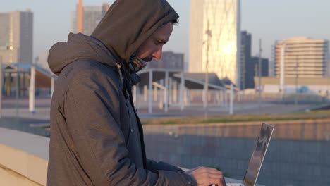 cold hooded male hacker quickly typing code on laptop overlooking downtown barcelona waterfront