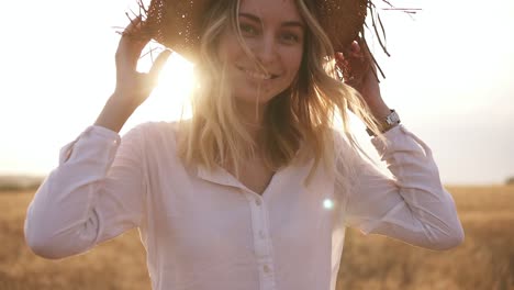 attractive, funny, smiling blonde woman in white shirt and straw hat posing while walking by wheat field on sunny summer day