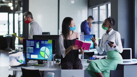 Diverse-female-office-colleagues-wearing-face-masks-discussing-at-modern-office