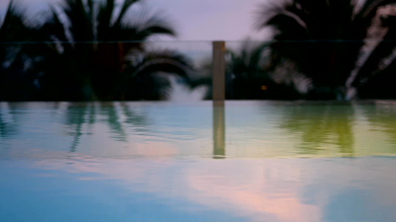 4K, close up of swaying coconut palm tree leaves or midrib from sea wind at evening time after sunset, loop. swimming pool at foreground with copy space. footage for chill leisure vacation background