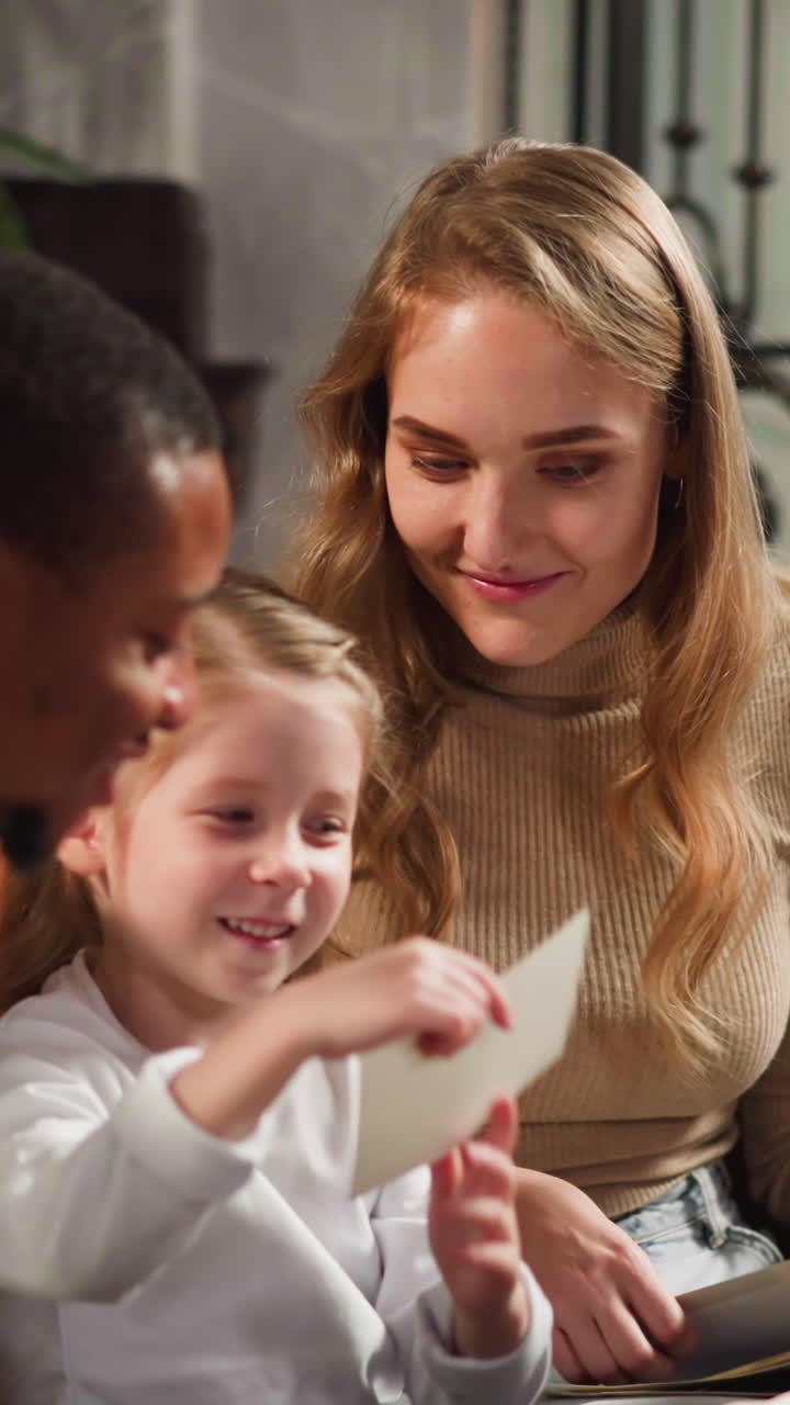 Smiling little girl shows vintage faded photos of relatives to black man guest enjoying family memories with blonde mother at home slow motion