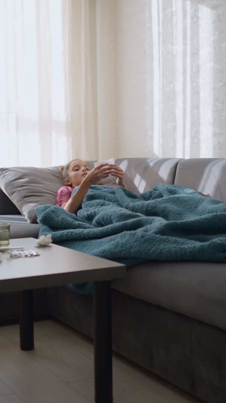 Unwell child lying on couch with tissue paper scattered around her, sick child covered in blanket resting in living room, surrounded by tissues, appearing tired and sick