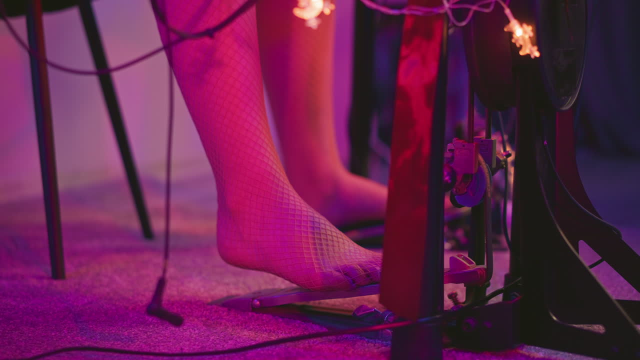 Leg view of barefoot woman in fishnet pantyhose stepping on kick pedal during electronic drum practice in softly lit studio with purple glow and visible wiring