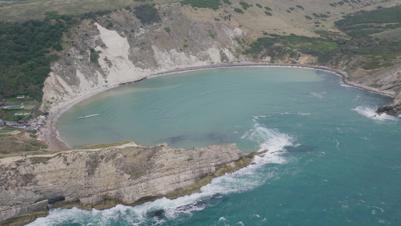 Suave toma aérea hacia atrás que revela Lulworth Cove en Dorset con su bahía curva, acantilados de piedra caliza, aguas turquesas y el escarpado paisaje de la Costa Jurásica