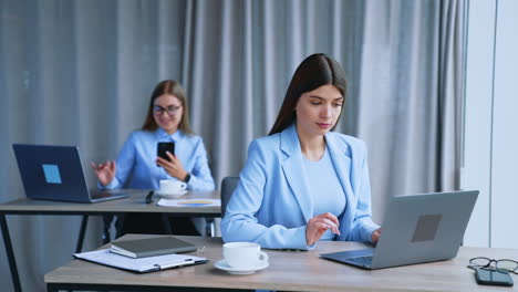 women work in the office sitting at the computers. brunette lady is excited and happy. blonde woman holding phone at backdrop in blur.