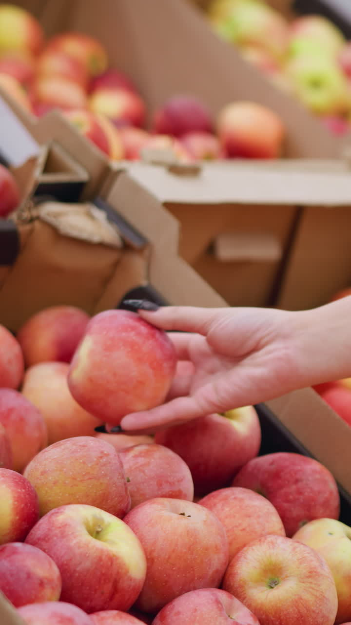 primer plano de una mujer seleccionando una manzana roja fresca de una pila de manzanas en una caja de cartón en una tienda de comestibles, mostrando la mano con uñas negras alcanzando la fruta