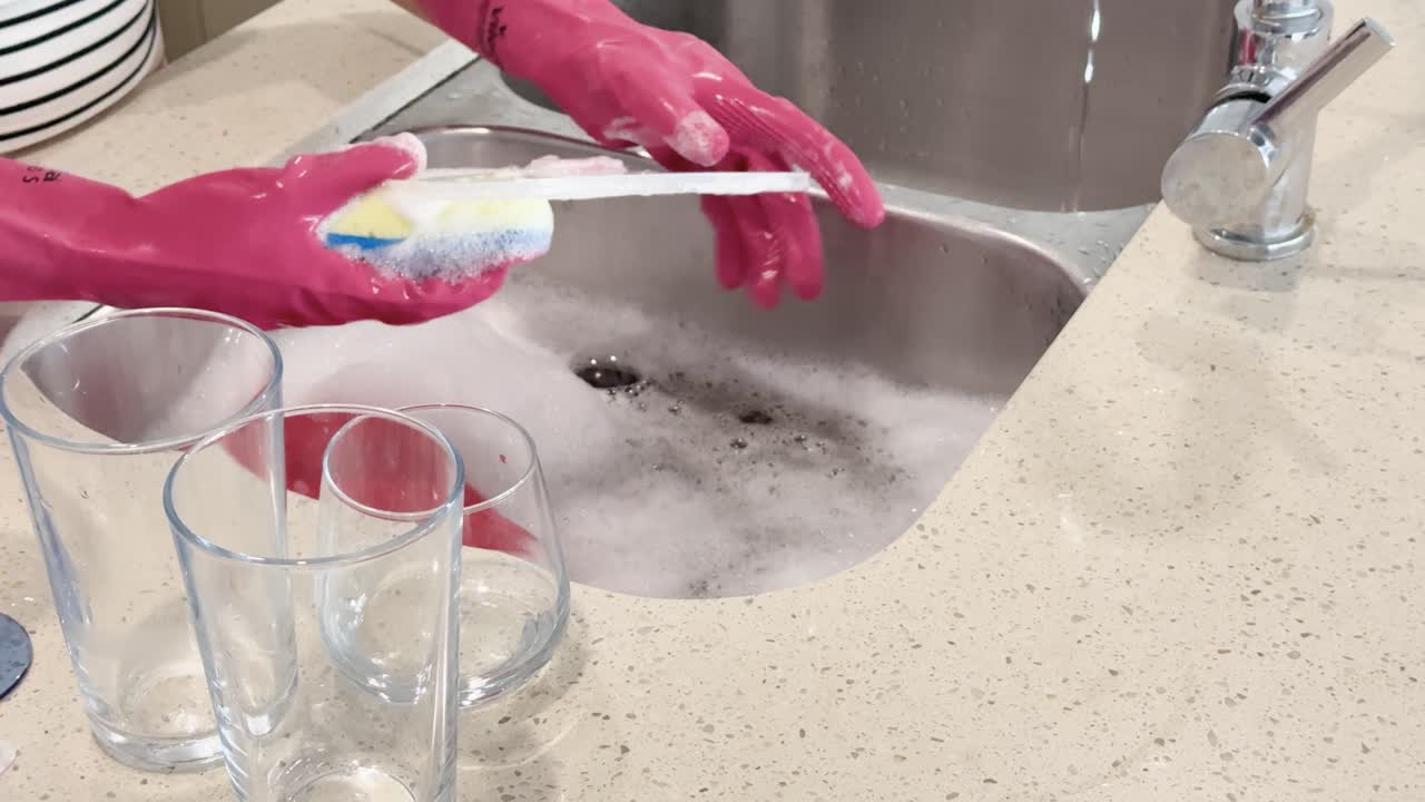 Person wearing pink gloves scrubs and rinses glass and plastic dishes in a stainless steel sink with soapy water under bright indoor lighting